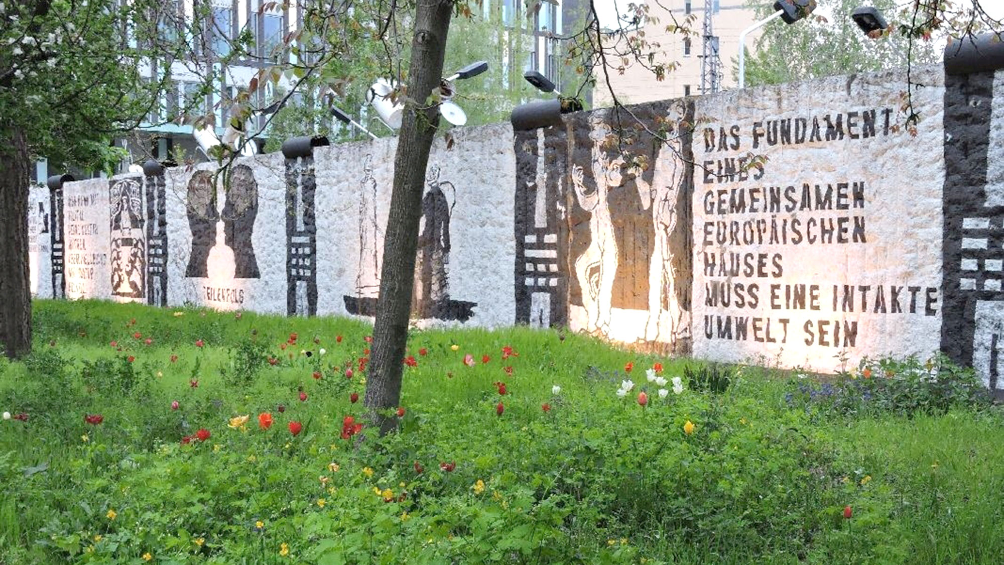 Blick auf das Parlament der Bäume - zu sehen ist ein Abschnitt der Berliner Mauer in einem begrünten Garten 