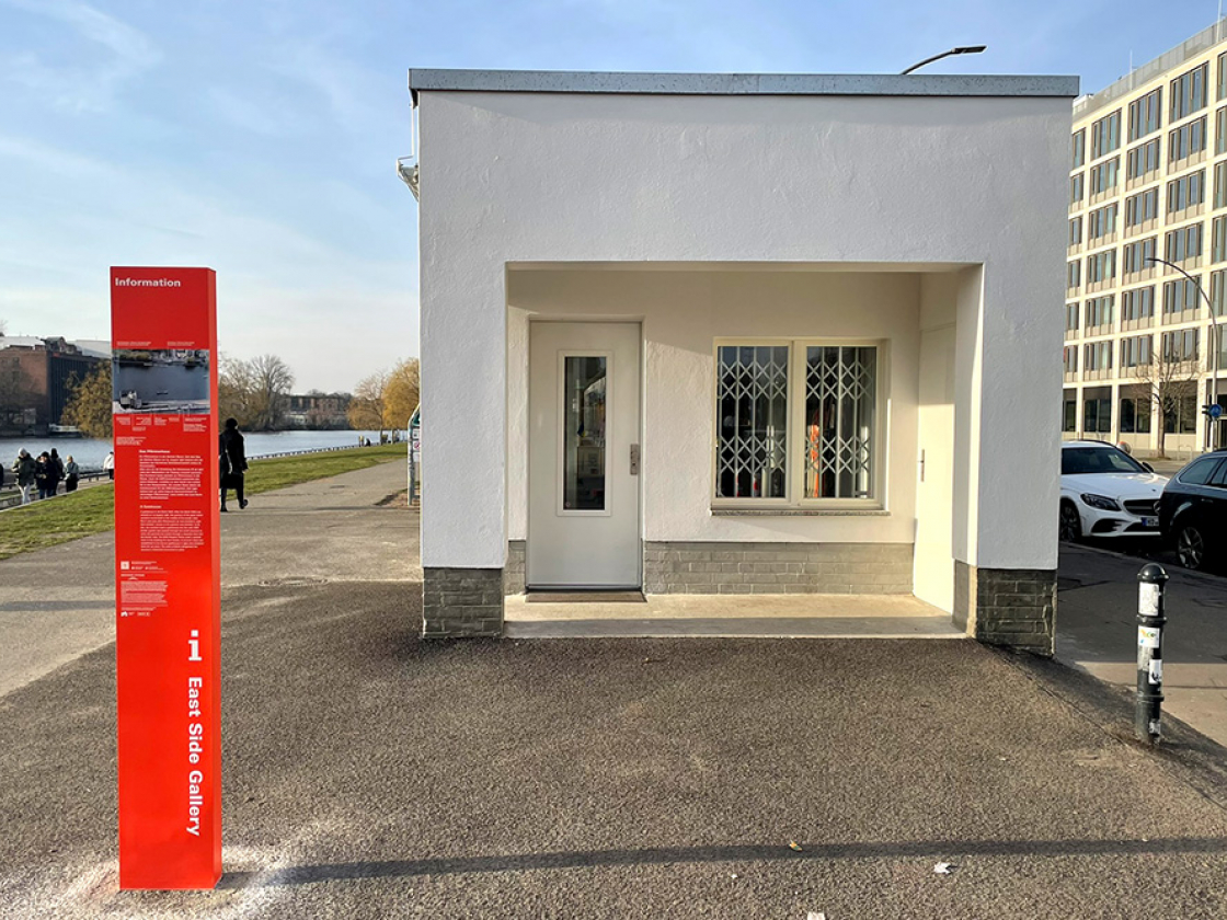 Man sieht den Eingangsbereich eines kleinen Hauses an der East Side Gallery und links eine orangene Stele mit Informationen.